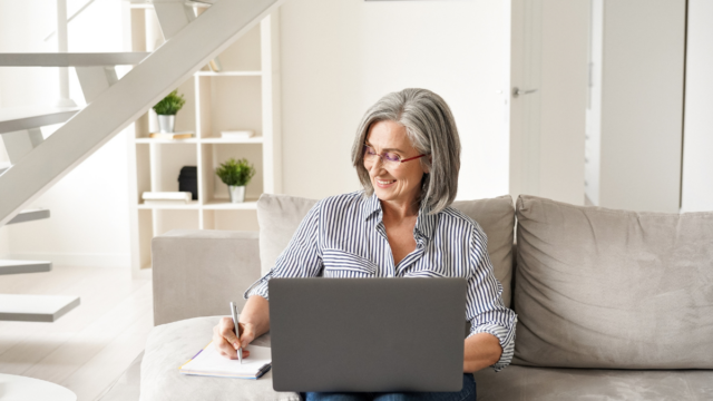 Assistante virtuelle caucasienne âgée, assise sur son canapé et travaillant sur son ordinateur portable tout en écrivant. 