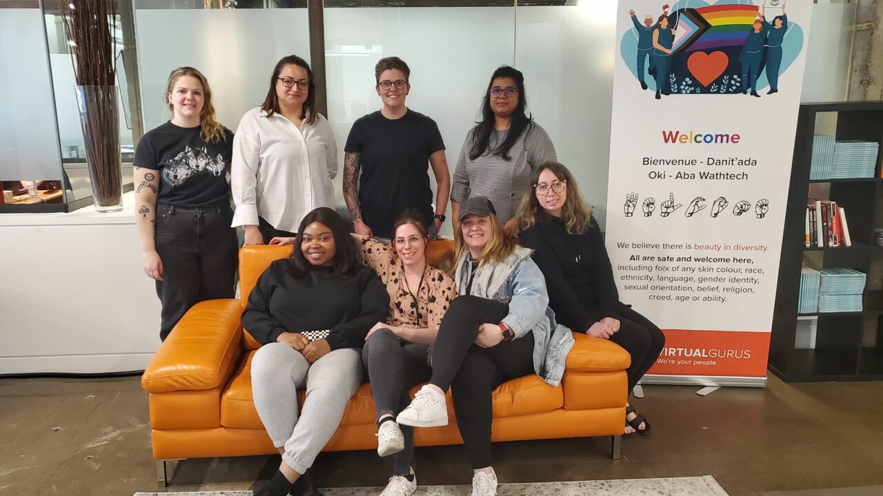 CSM team photo with staffers sitting on the orange couch or standing behind it at the Virtual Gurus office with a Welcome stand up sign beside them.