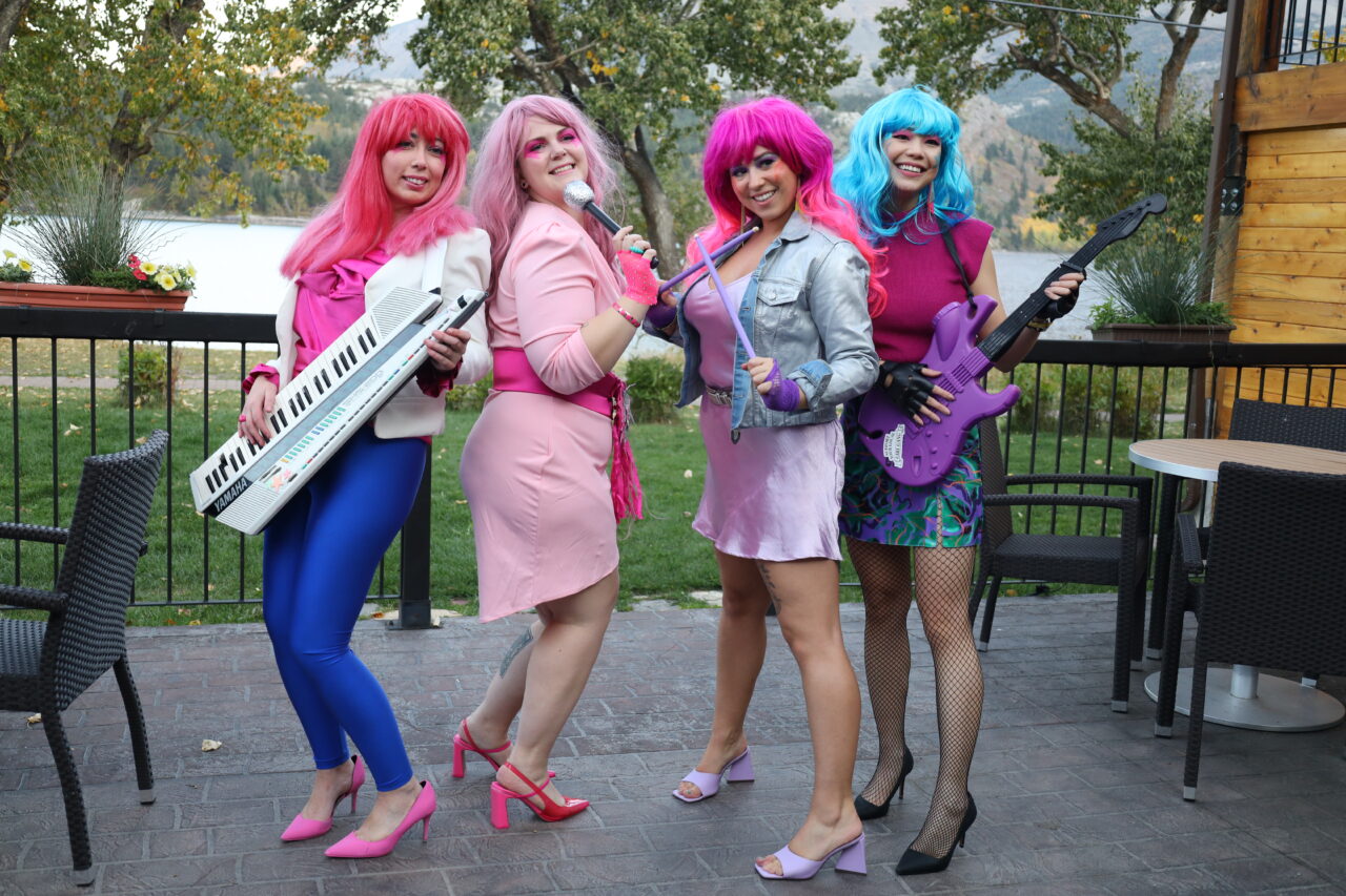 A group of Virtual Gurus staff members dressed as vibrant rockstars taking a fun group photo together on an outdoor patio with a lake in the background.