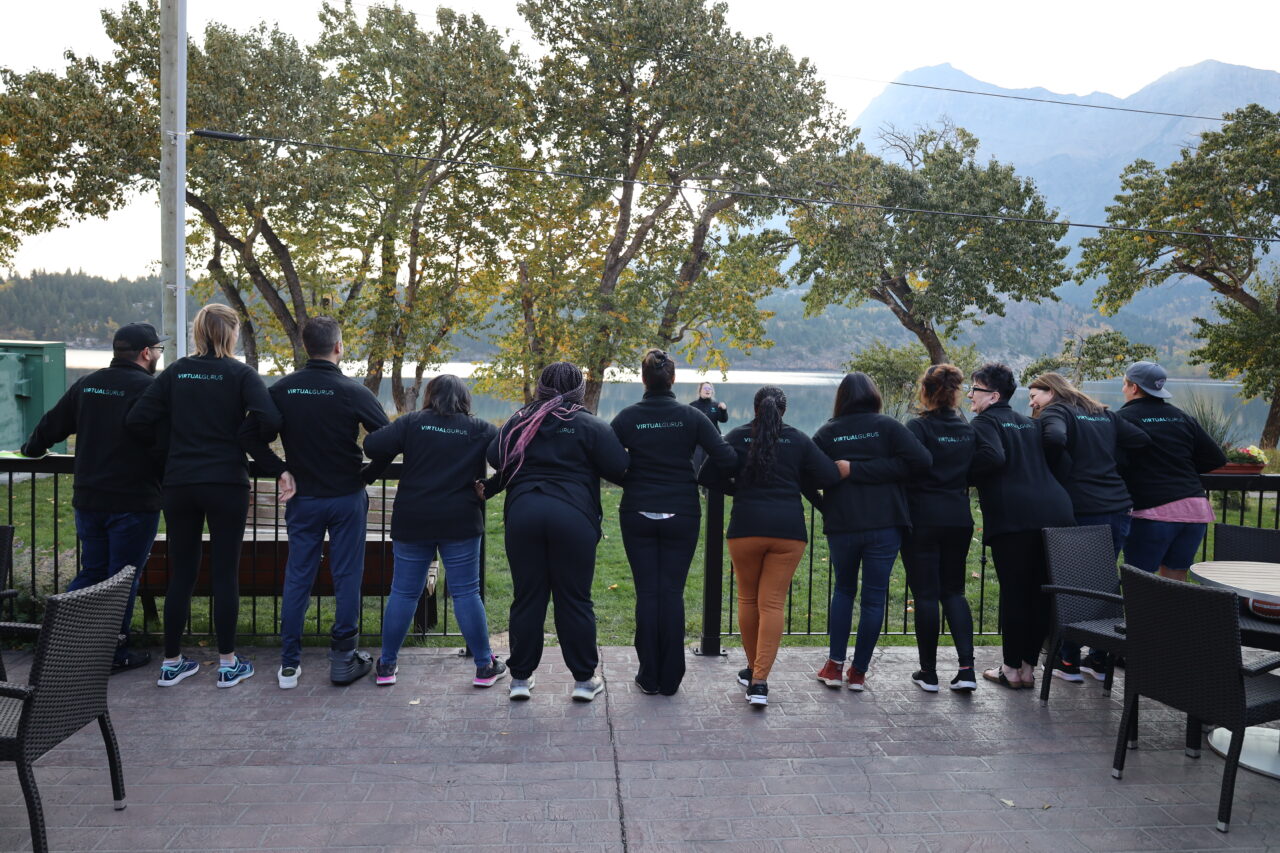 A group of Virtual Gurus staffers all lined up against a railing on a patio with their backs to the camera wearing matching black company jackets with a lake and mountains in the background.