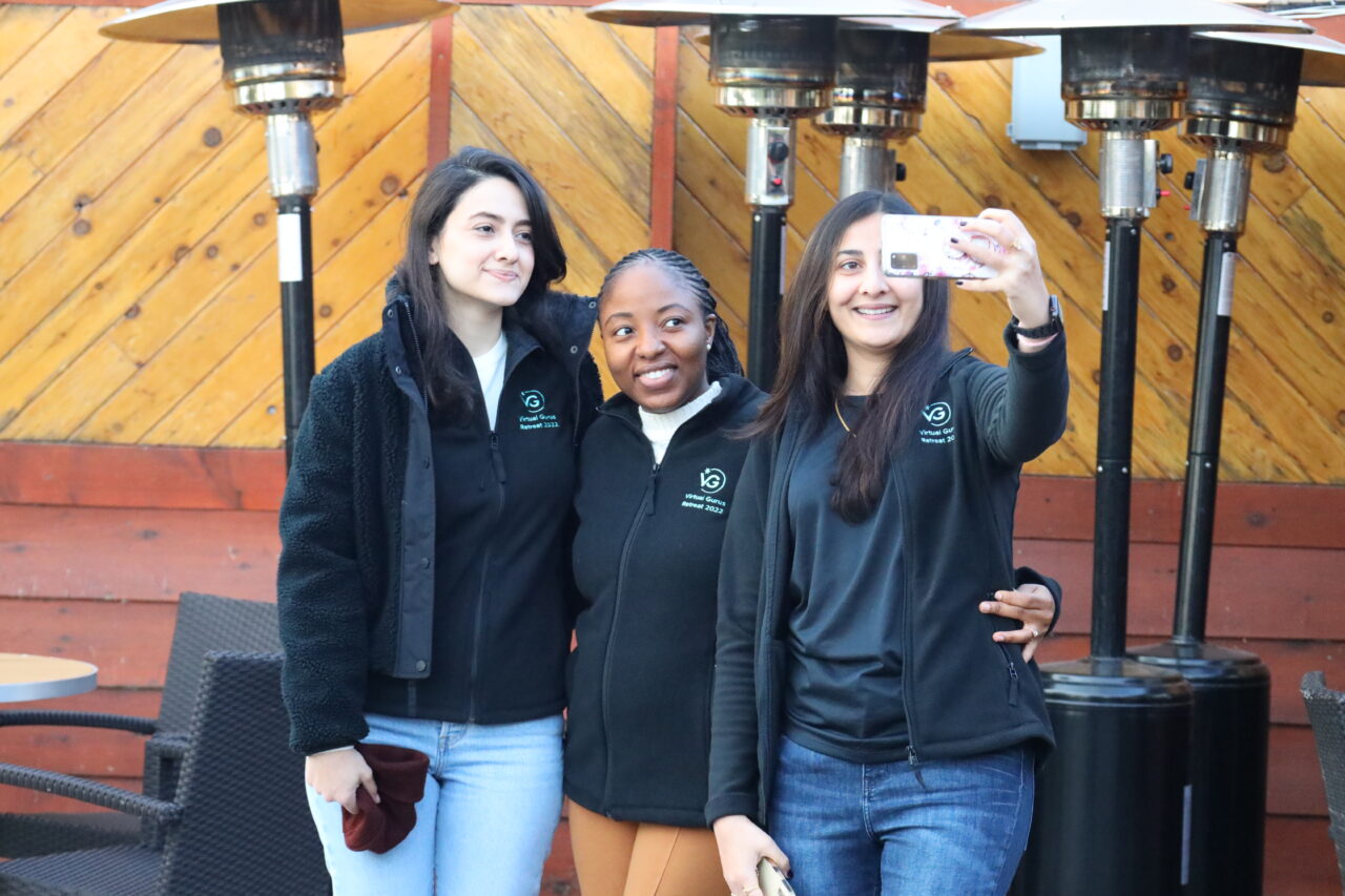 Three beautiful Virtual Gurus staff members, all wearing matching black company jackets, taking a selfie on a patio.