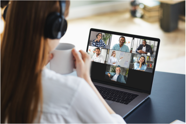 A blurred out woman with long brown hair wearing headphones sitting while holding a white mug on a laptop with attendees of a virtual meeting.