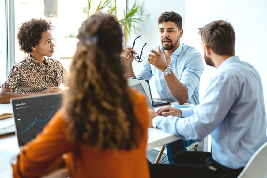 A Black man holding glasses is explaining something to a group of diverse staff at a boardroom table.