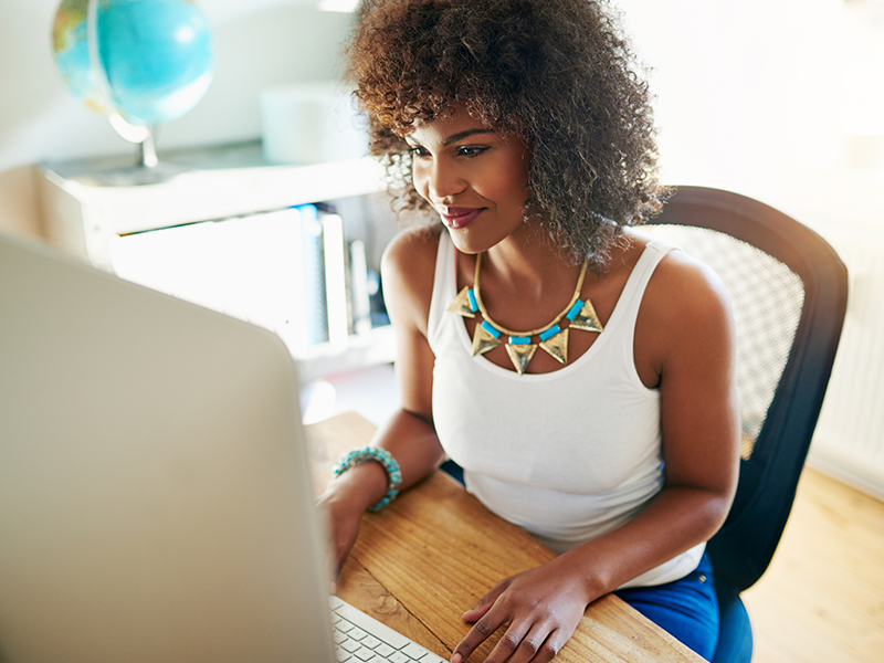Looking down at an African American woman as she sits at her desk smiling after hiring a virtual assistant. She's wearing a white tank top and bold yellow and blue necklace.