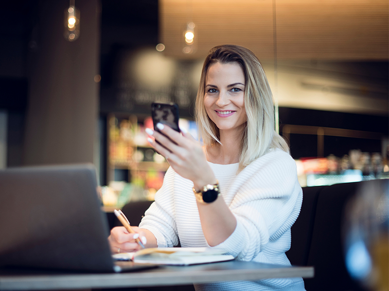 Caucasian woman with blonde hair is holding her phone while sitting in front of a laptop and writing on a notepad.