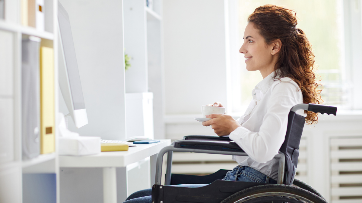 sideview of woman sitting in her wheelchair, holding coffee and smiling at computer screen
