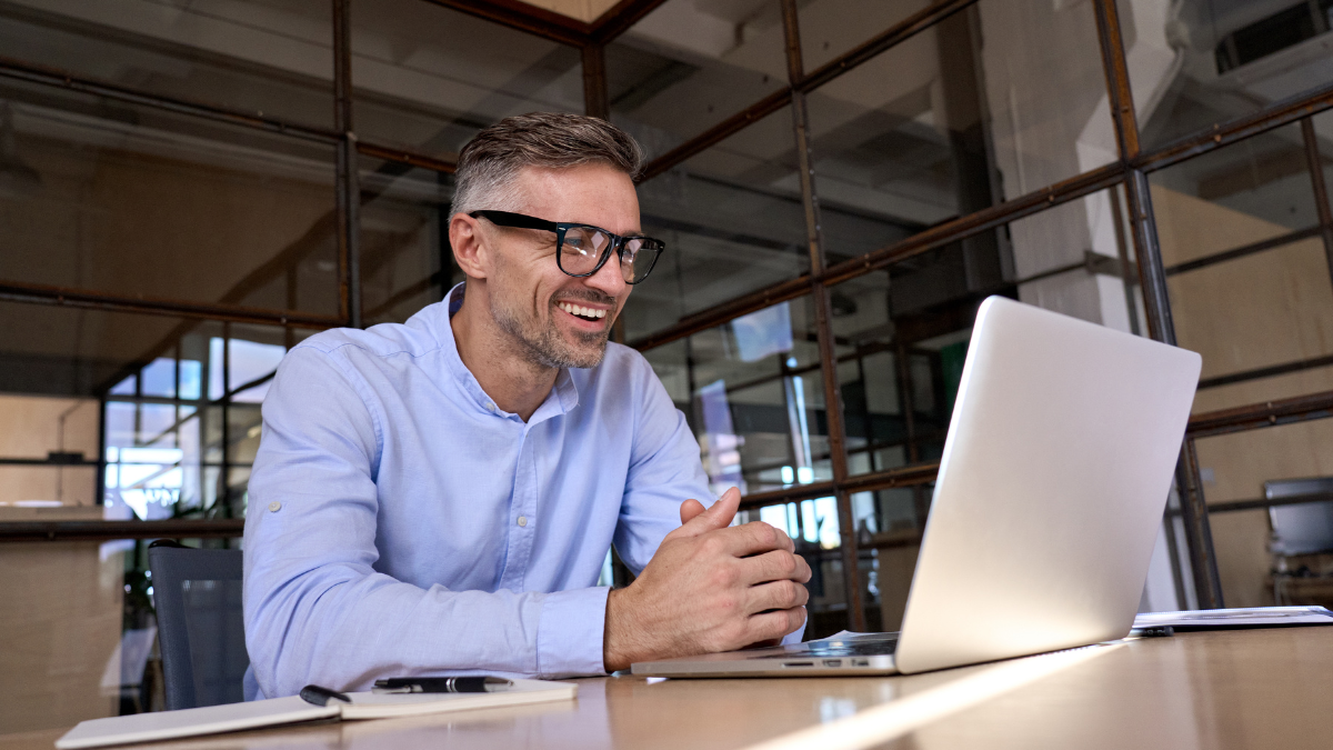 Homme caucasien portant des lunettes à monture noire, chemise bleue, assis à son bureau et souriant aux solutions économiques d'embauche d'un AV sur son écran d'ordinateur.