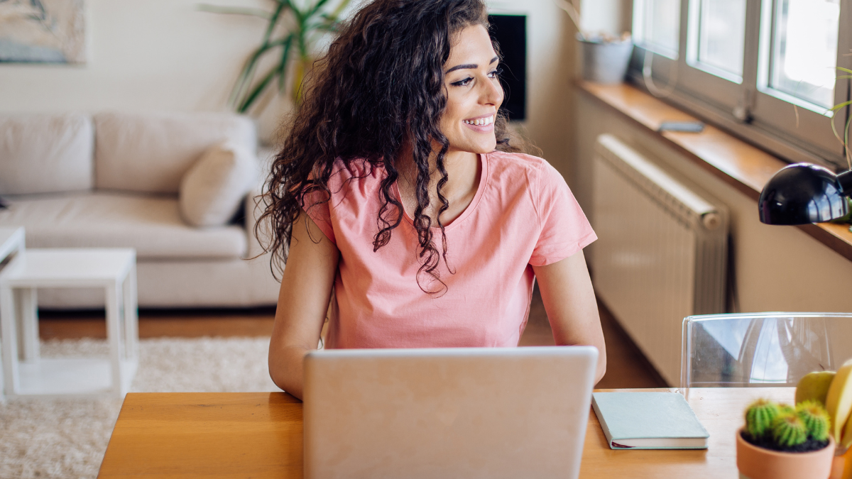Caucasian women with curly brown hair, wearing a pink shirt, sitting at her home office desk as a virtual assistant, in front of her laptop and looking out the window smiling