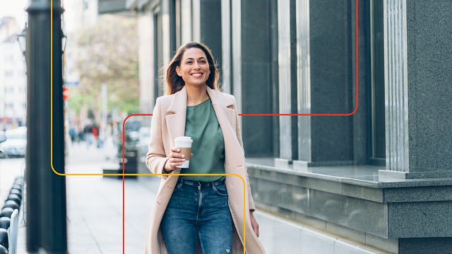 Caucasian woman, long blond hair, smiling, wearing jeans and long business coat, walking outside with a coffee in hand.