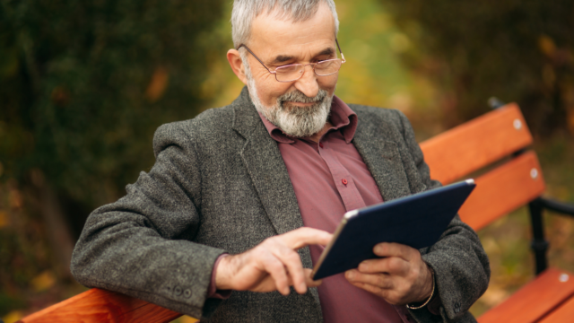Older man taking a work break and sitting on a park bench and smiling while scrolling on his tablet.