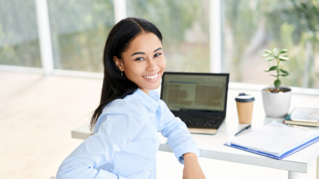 Woman of color, long black hair, virtual assistant, sitting in front of laptop, turning back and smiling.