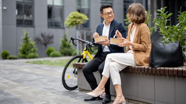 Asian man sitting with Caucasian woman outside, eating lunch while they strengthen their workplace mental health