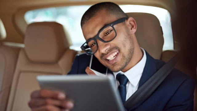 Black man wearing black glasses and a suit, sittin gin car and smiling while looking at tablet