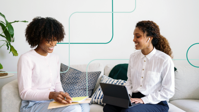 Two Black woman sitting on a couch and smiling while building their virtual assistant into their AI strategy